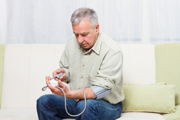 Senior man is sitting on sofa at home and  measuring blood pressure.