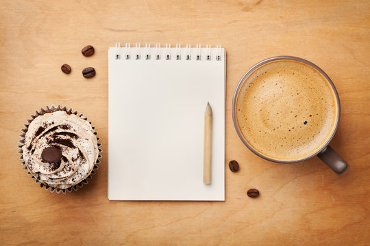 Coffee mug with cupcake, notebook and pencil on rustic table from above, cozy and sweet breakfast, good morning or have a nice day concept