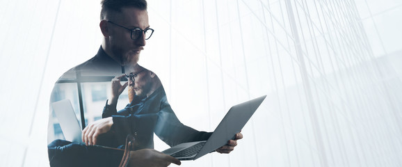 Double exposure bearded businessman wearing black shirt and glasses,holding contemporary notebook hands. Portrait young banker using laptop in modern office, skyscraper background. Wide
