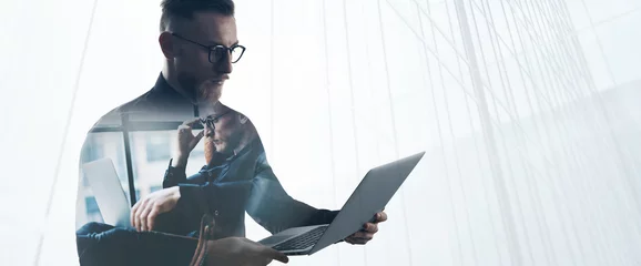 Double exposure bearded businessman wearing black shirt and glasses,holding contemporary notebook hands. Portrait young banker using laptop in modern office, skyscraper background. Wide © SFIO CRACHO