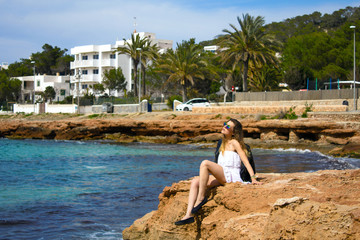 Young classy girl sitting on rocky coast of the ibiza island during hot summer