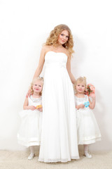 Fototapeta na wymiar Beautiful blond mom with two young daughters blondes in white dresses on a white background. Beautiful mother and daughters in studio