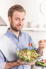 Fototapeta na wymiar Happy smiling man eating fresh vegetable salad in the kitchen