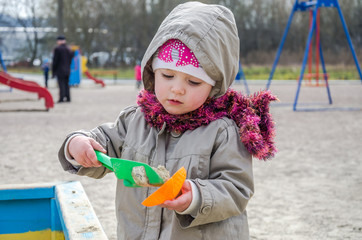 Little lovely girl baby playing in the sandbox on the playground with a shovel and bucket digging a hole, dressed in a raincoat with a hood, scarf and hat