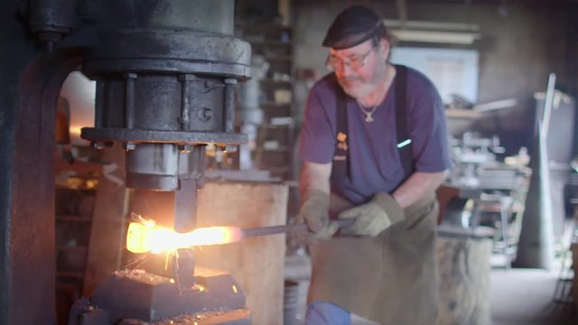 Blacksmith using a large industrial hammer to work on a hot iron bar.  Medium shot with some metal sparks.  Originally recorded hand-held in 4K and cropped.