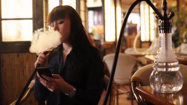 Beautiful young woman inhaling hookah and using phone. girl smoking shisha in cafe. Warm light.