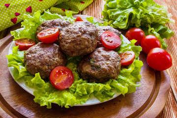 Roasted beef cutlets, green salad and small tomatoes on white plate. Wooden background.
