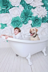 Naklejka premium little cute 4-years boy with funny face in bathrobe with his dog labrador retriever sit in the bath and smile with white and turquoise flowers on the background