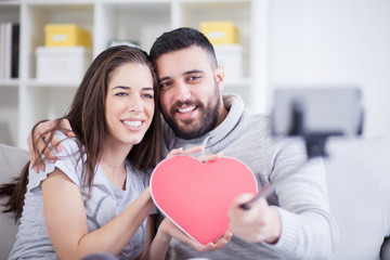 Happy young beautiful couple taking photo with smart phone on selfie stick. Young couple having fun in the living room