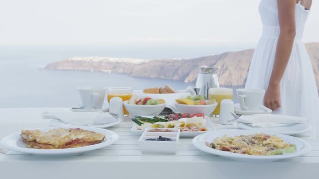 Delicious breakfast by the sea. Perfect breakfast table outdoors. Elegant tourist woman in white dress walking by table. Caldera view on Santorini, Greece, Europe.