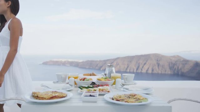 Breakfast table on terrace with woman walking by. Delicious food served at resort by the sea and mountain. Tourist on vacation travel holidays in Santorini with view of Caldera.