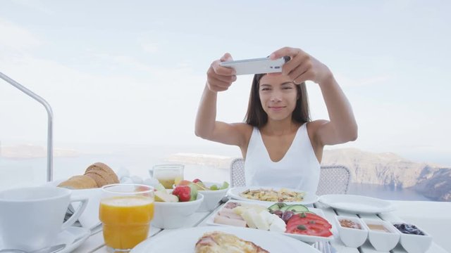 Tourist woman photographing breakfast using mobile smart phone app.  Girl taking picture of food on luxury travel vacation for social media. Beautiful female in resort in Santorini, Greece, Europe.