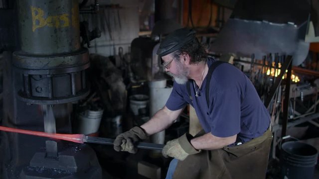 Blacksmith working hot iron bar with a large industrial hammer.  High viewpoint, medium shot, originally recorded in 4K with hand-held camera.