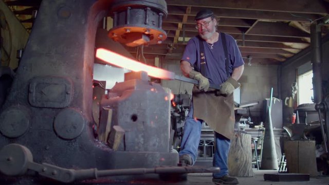 Blacksmith working hot metal with a large industrial hammer.  Low angle wide shot, originally recorded in 4K at 60fps.