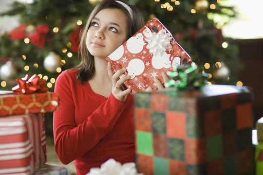 A girl by a Christmas tree shaking a present trying to guess what it is.