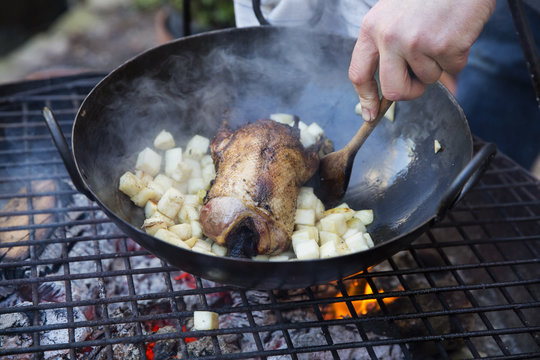 An outdoor cookout, a game bird in a pan surrounded by mirepoix of vegetables above a glowing fire. 