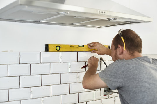 A man working in a new kitchen, a tiler applying tiles to the wall behind the cooker. 