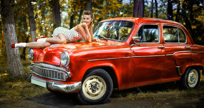 Pin-up girl in white dress on the hood of red retro car on a background of green forest