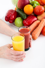 Mature female hands holding two glasses with red and yellow fresh juices, fruits and vegetables are on white table