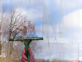 Hand in pink protective glove washing and cleaning window with professionally squeegee on background of cloudy sky. Early spring windows cleaning. Maid cleans window.