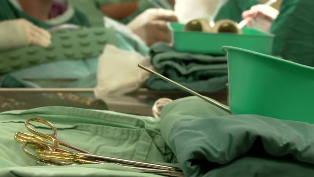 Hospital. Medicine. Sterile surgical instruments in the front and surgeons team performing operation in hospital operating room at the background. Hands close up. Shot 12