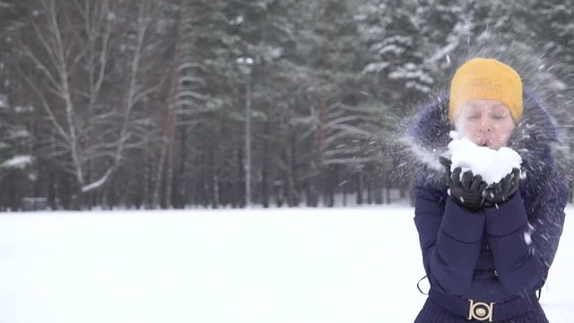 Playful woman with yellow hat blow fluffy snow and skier man on background in winter park. Static shot. 4K
