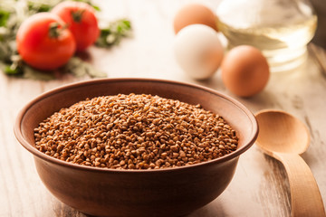 Deep dish with raw buckwheat on a wooden table on a background of the tomato and eggs
