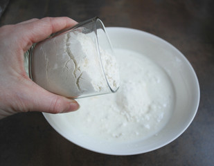 Female hand holding a transparent glass with the flour over the dough. Preparation for cooking. 