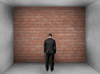 Businessman with bowed head stands front of a brick wall in interior. 