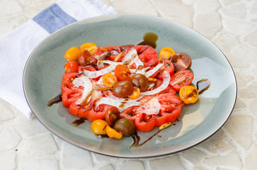 A vibrant healthy mixed tomato, mozzarella, pine nut and balsamic vinegar salad, served on a rustic plate on a tile table.