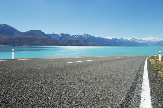 Empty Asphalt Road Near Water And Snow Mountains