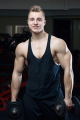Fototapeta na wymiar Smiling muscular man posing in a black tank top and black shorts with dumbbells in the gym