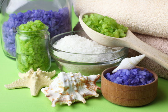 sea salt poured into a glass jar and bowl with a wooden spoon and fluffy towel with sea shells on a green background