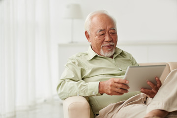 Senior Vietnamese man sitting on sofa and reading new on his tablet computer - Powered by Adobe