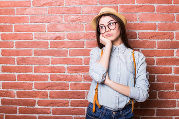 Portrait of beautiful girl in casual clothes and hat showing bored, looking in camera and smiling while standing against red brick wall