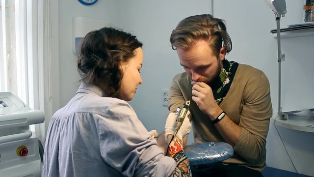 A female tattoo artist in gloves is making the picture of a monkey on a young man's hand. The procedure takes place in the office using a tatoo machine.