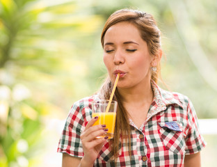 Young woman in square pattern shirt enjoying a yellow juice with eyes closed, garden environment