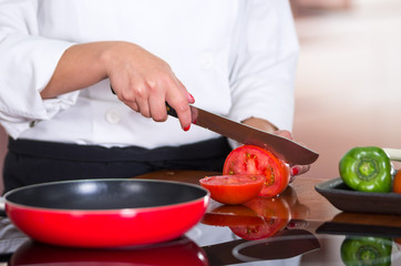 Closeup chef cutting tomato on wooden surface with red skillet in front