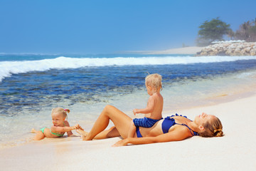 Happy family - mother, baby son and daughter lying on sand beach and look at sea surf after swimming in clear water. Active parents and people outdoor activity on tropical summer vacations with child