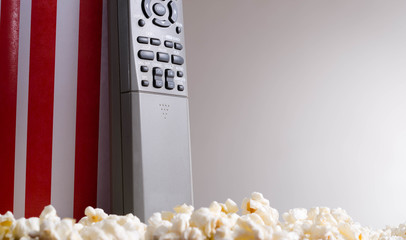Closeup red white striped container standing up with popcorn lying around, remote control leaning on box, low angle, grey background