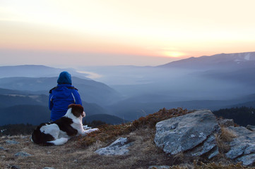 Young woman with dog watching sunrise high in the mountain - obrazy, fototapety, plakaty