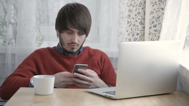 Happy Young Man Freelancer Working with Modern Laptop and Phone in Cafe taking Cup of Coffee