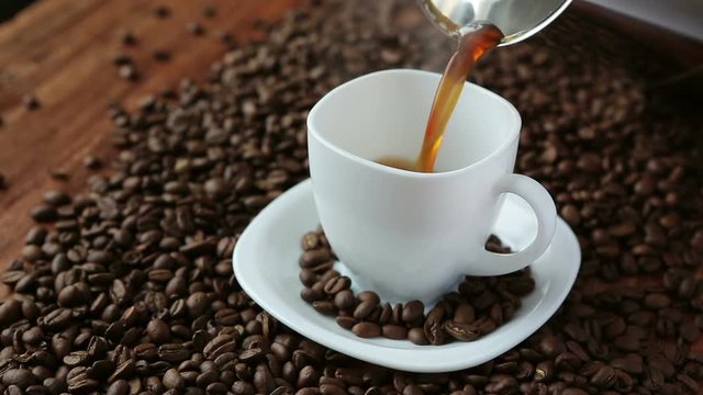 Pouring flavored coffee from coffee pot in white cup surrounded by coffee beans on wooden table. Close up view