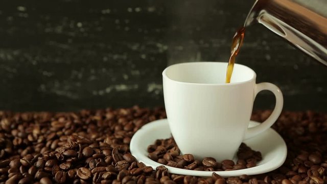 Pouring coffee from coffee pot in white cup surrounded by coffee beans on dark background