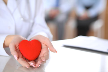 Female doctor with stethoscope holding heart.  Patients couple sitting in the background