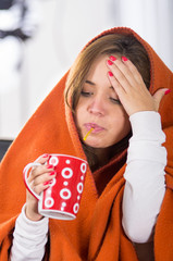 Brunette sitting down under orange blanket, holding red cup in hand, sick with flu concept