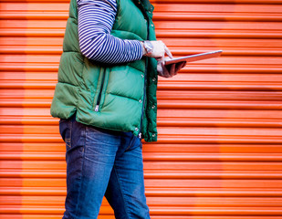 Young man using a tablet while walking in front of an orange self storage garage door.  Dressed casually. Jeans, Vest. Urban life style, technology, business, self storage  and fashion concept 