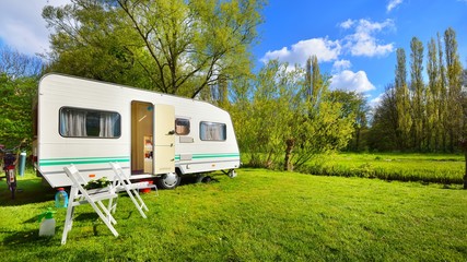 White caravan trailer on a green lawn in a camping site. Sunny day. Spring landscape. Europe. Lifestyle, travel, ecotourism, road trip, journey, vacations, recreation, transportation, RV, motorhome - obrazy, fototapety, plakaty