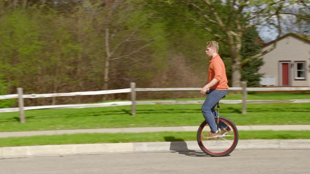 Young man rides unicycle around Mid West suburban street, then dismounts by stepping off.  Wide shot recorded in 4K on a spring day in the American Mid West.