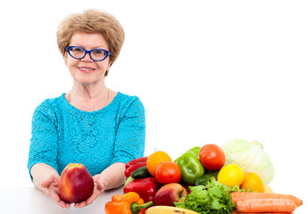 Happy elder Caucasian woman stretching hands with big red apple, vegetables and fruits are on table, isolated on white background